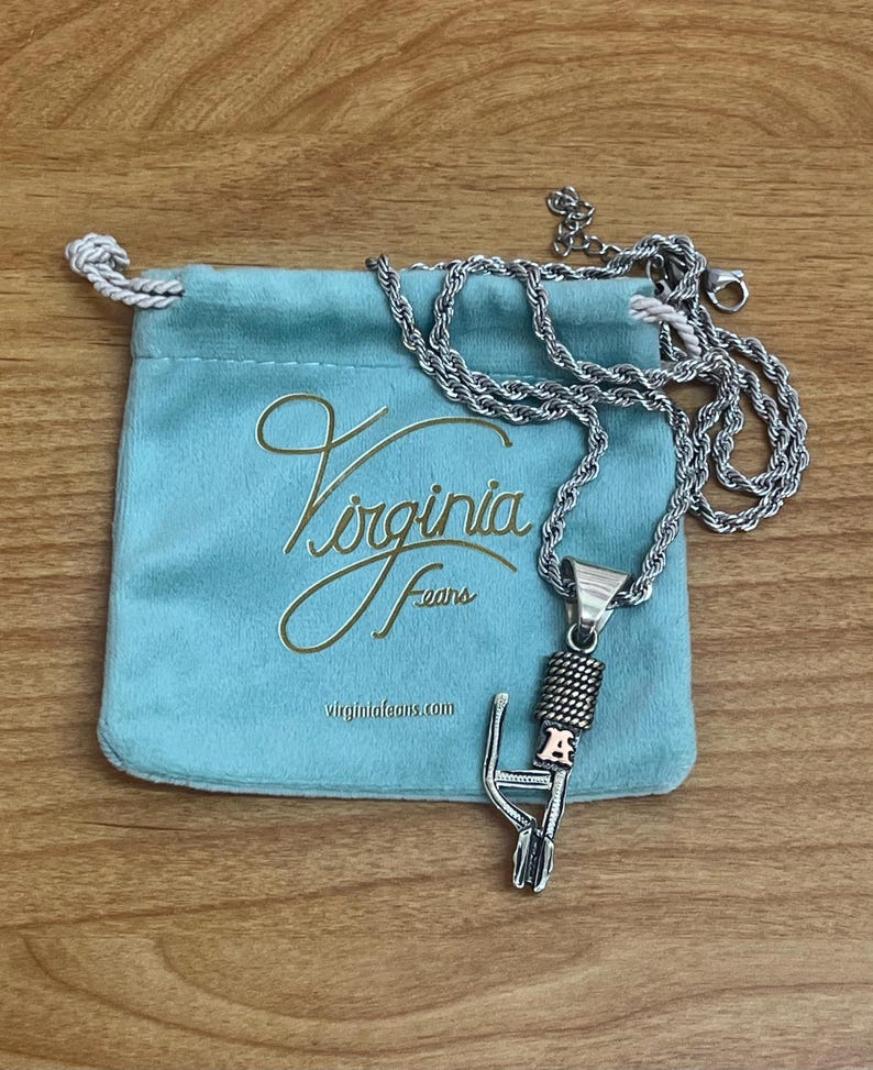 Puede incluir: Un collar con colgante de micr&oacute;fono plateado con una cadena de cuerda descansa sobre una bolsa de terciopelo azul claro. La bolsa tiene letras doradas que dicen "Virginia Feans" y la direcci&oacute;n del sitio web. El collar est&aacute; sobre una superficie de madera.