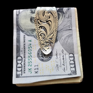 May include: A silver money clip with an ornate floral design holds a stack of U.S. $100 bills. The clip has a detailed pattern and the text "Jorgensen" is visible. The bills are crisp and new.