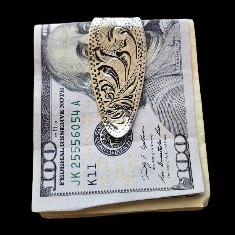 May include: A silver money clip with an ornate floral design holds a stack of U.S. $100 bills. The clip has a detailed pattern and the text "Jorgensen" is visible. The bills are crisp and new.