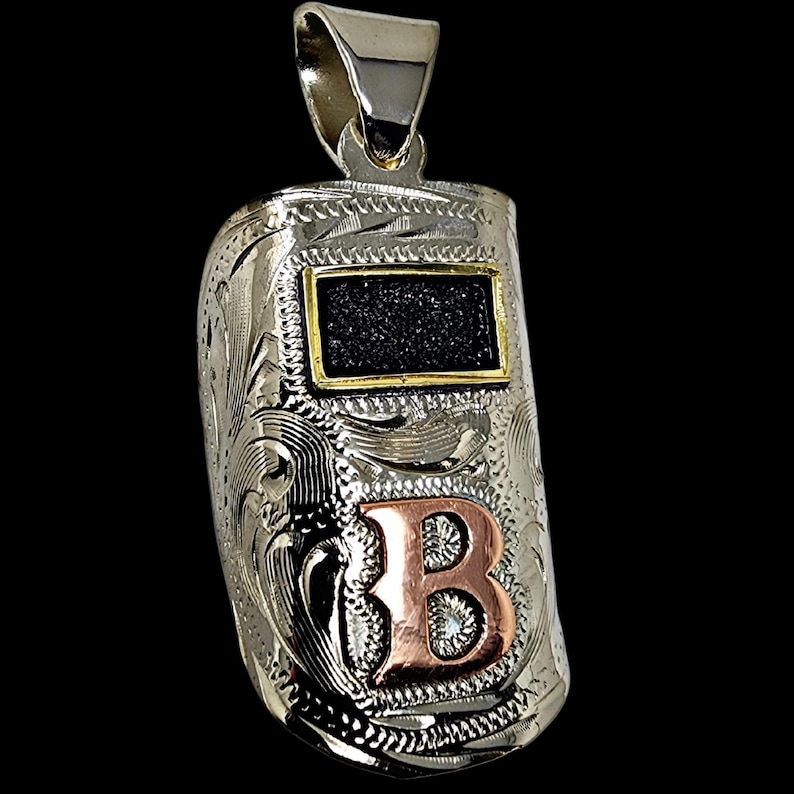 May include: Silver pendant shaped like a welding mask, featuring intricate floral engraving. The mask has a black rectangle with a gold border and a copper-colored letter "B". The pendant hangs from a silver bail.