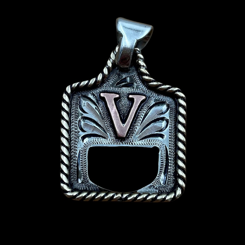 May include: Silver pendant with a decorative rope border and a copper-coloured letter "V" in the centre. The pendant has an ornate, engraved design and a bail for a chain. The background is black.