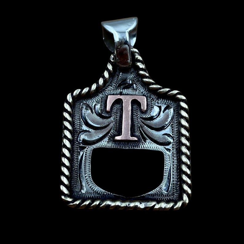 May include: Silver pendant with a decorative rope border and a copper-coloured letter "T" in the centre. The pendant has an engraved design and a dark background.