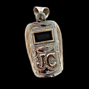 May include: Silver pendant with a rectangular black center framed in gold. The pendant features engraved floral patterns and gold-colored initials "JC". The bail is silver. The pendant is likely made of metal.