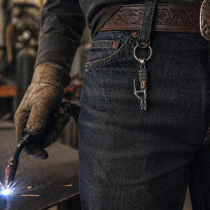 Puede incluir: Un llavero de metal plateado con la letra "J" est&aacute; sujeto a una trabilla del cintur&oacute;n. El llavero cuelga de un cintur&oacute;n de cuero marr&oacute;n con un dise&ntilde;o decorativo. La persona lleva vaqueros azules y una camisa de manga larga gris oscuro. Una antorcha de soldadura est&aacute; en la mano de la persona.