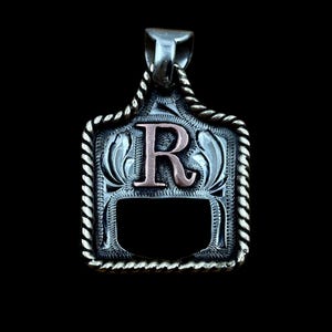 May include: Silver pendant with a decorative border and a copper-coloured letter "R" in the centre. The pendant has a rectangular shape with a rope-like edge and a bail for a chain. The background is black.
