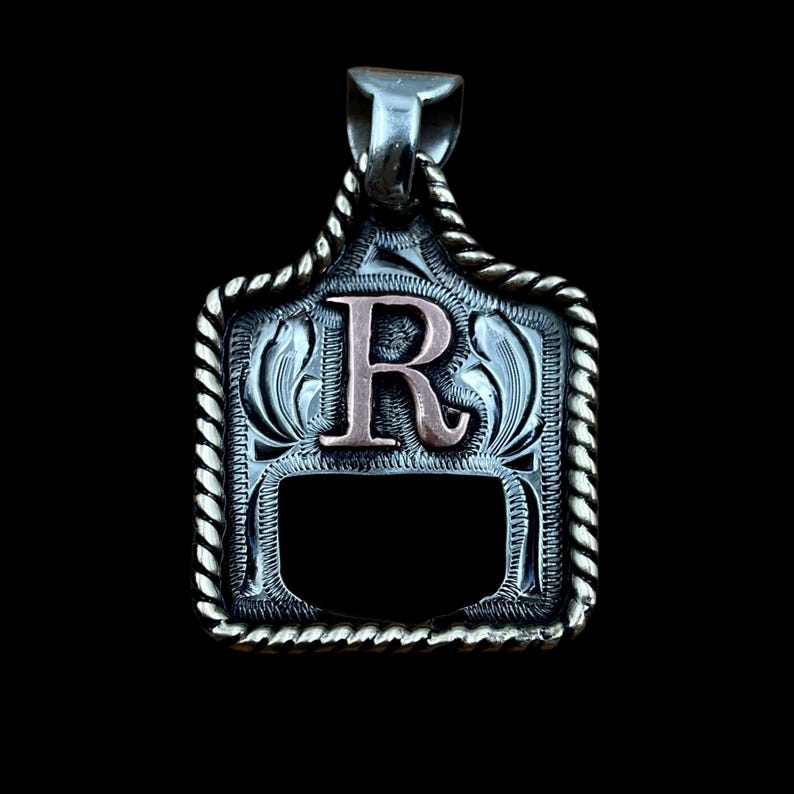 May include: Silver pendant with a decorative border and a copper-coloured letter "R" in the centre. The pendant has a rectangular shape with a rope-like edge and a bail for a chain. The background is black.