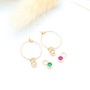 Peut inclure: Deux boucles d'oreilles créoles en or avec de petits pendentifs qui en pendent. Les pendentifs sont ronds et présentent une pierre transparente, verte et rose.