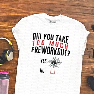 May include: White t-shirt with the text "Did you take too much preworkout?" and a checkbox with the options "Yes" and "No". The text is in red and the checkbox is black. The shirt is on a wooden surface with weights and other fitness equipment.