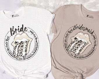 Op de afbeelding: Twee t-shirts, één witte en één beige, met een distressed design. Het witte shirt zegt "Bride" en het beige shirt zegt "Bridesmaid". Beide hebben een cirkelvormige afbeelding met een tongontwerp en de zin "We Only Roll with the Best".
