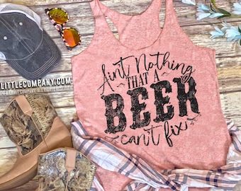 May include: Pink tank top with the text "Ain't nothing that a beer can't fix" in black. The tank top is laying on a wooden surface with a pair of brown cowboy boots, a plaid shirt, and a denim skirt.