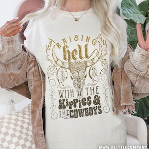 May include: A white t-shirt with a brown graphic design featuring a skull with horns and the text "Raising hell with the hippies & the cowboys".