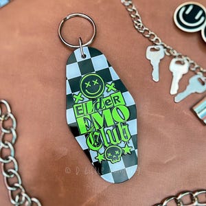 May include: A black and white checkered keychain with the words "Elder Emo Club" in neon green. The keychain has a smiley face with crossed-out eyes and a skull with stars. A silver chain and keys are in the background.