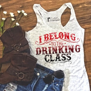 May include: A casual outfit featuring a gray tank top with the text "I BELONG TO THE DRINKING CLASS", denim shorts, brown cowboy boots, and sunglasses. The tank top has a racerback design. The shorts are distressed. The boots have buckles.