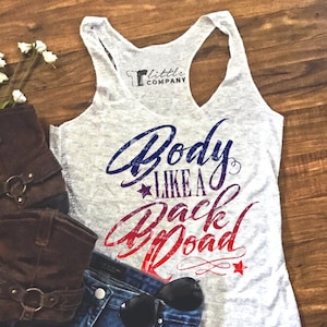 May include: A gray tank top with the text "Body Like A Back Road" in red, white, and blue. The text is in a distressed font. The tank top is laying on a wooden surface with a pair of brown boots, denim shorts, and sunglasses.