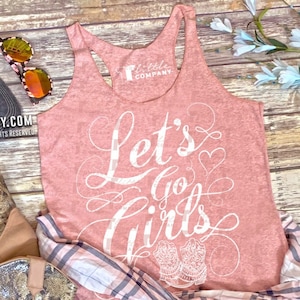 May include: A blush pink tank top with the text "Let's Go Girls" in white script. The text is surrounded by a heart and a pair of stylized cowboy boots. The tank top is laid out on a wooden surface with a plaid shirt, a pair of cowboy boots, and a baseball cap.