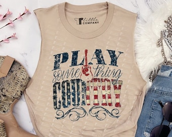 May include: A beige sleeveless top with the text "PLAY something COUNTRY" in blue and red, paired with denim shorts featuring white star patterns. The image also includes snakeskin-patterned boots and a silver necklace.