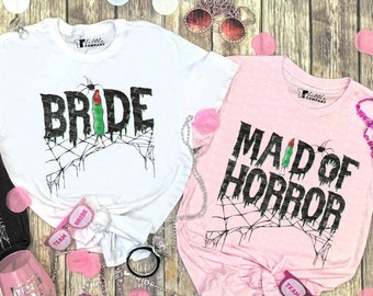 May include: Three black, white, and pink t-shirts with a horror-themed bridal party design. The shirts say "Bride", "Bridesmaid", and "Maid of Horror".