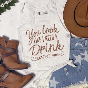 May include: A gray tank top with the text "You look like I need a drink" in brown lettering. The tank top is styled with denim shorts, a brown hat, and brown ankle boots.