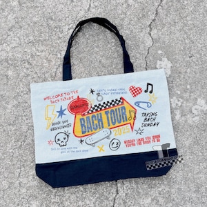 May include: A light blue tote bag with a navy blue bottom and handles. The bag features colorful text and graphics, including "BACH TOUR 2025", a skateboard, and the phrase "Nobody likes you when you're the bride to be."