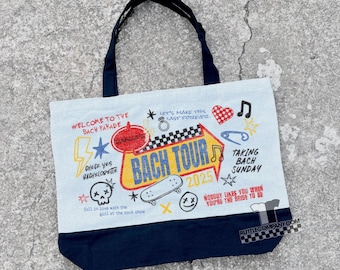Può includere: Una borsa tote azzurra con fondo e manici blu navy. La borsa presenta testo e grafica colorati, tra cui "BACH TOUR 2025", uno skateboard e la frase "Nobody likes you when you're the bride to be."