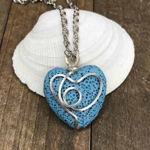 May include: A blue lava rock heart pendant necklace with a silver wire design. The necklace is on a silver chain and is resting on a white seashell.