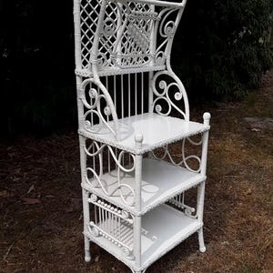 May include: White wicker etagere with three shelves and a decorative top. The etagere features intricate woven patterns, scrollwork, and a lattice back. The shelves are rectangular and the piece stands approximately 60 inches tall.