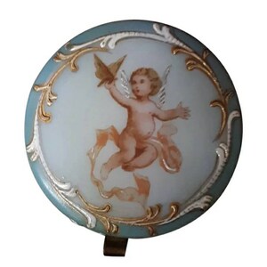 May include: A round, blue and white porcelain trinket box with a gold trim. The lid features a painted image of a cherub with wings holding a bird.