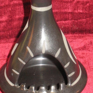 May include: Black ceramic ashtray with a white design. The ashtray is shaped like a small house with a rounded top and a wide opening in the front. The ashtray has a decorative rim around the opening.