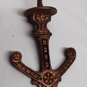May include: A bronze-colored metal wall hook shaped like an anchor. The hook features intricate embossed floral and geometric patterns along its arms and central shaft. A threaded screw extends from the top circular mounting plate for installation.