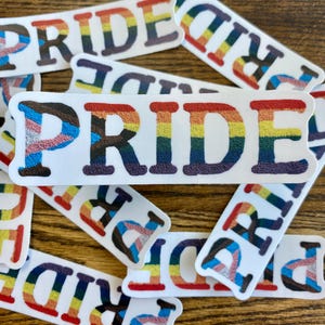 May include: A close-up of several stickers with the word "PRIDE" written in rainbow colors. The stickers are arranged in a pile on a wooden surface.