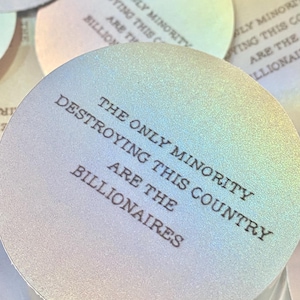 May include: Close-up of several iridescent, circular stickers. The text on the stickers reads: "THE ONLY MINORITY DESTROYING THIS COUNTRY ARE THE BILLIONAIRES." The stickers have a subtle, shimmering effect with a gradient of colours.