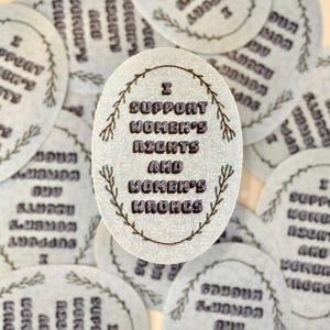 Puede incluir: Primer plano de una pegatina con fondo blanco y un diseño de corona verde. La pegatina dice "Apoyo los derechos de las mujeres y los errores de las mujeres".