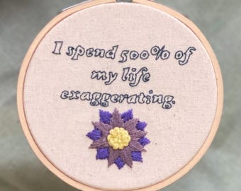 Könnte beinhalten: Gesticktes Stickrahmen-Kunstwerk mit dem Text "I spend 500% of my life exaggerating." Unter dem Text ist eine lila-gelbe Blume gestickt. Die Stickerei befindet sich in einem Holzreifen.
