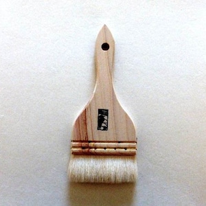 May include: A natural wood-handled paintbrush with a wide, rectangular head and a pointed tip. The brush features a light-colored wooden handle with a small, black rectangular stamp. The bristles are white and appear soft.