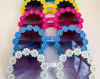 May include: Five pairs of sunglasses with daisy flower accents. The sunglasses are in different colors, including green, yellow, pink, blue, and white.
