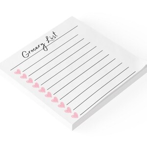 May include: A white notepad with the words "Grocery List" in cursive script. Pink heart shapes are drawn next to the lines. The notepad is a stack of white paper.