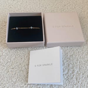 Puede incluir: Una pulsera de cadena dorada con dos piedras preciosas azul claro, presentada en una caja negra abierta. La caja est&aacute; dentro de una caja rosa claro y una tarjeta plegada, todas con el texto "S FOR SPARKLE".