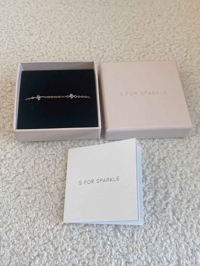 Puede incluir: Una pulsera de cadena dorada con dos piedras preciosas azul claro, presentada en una caja negra abierta. La caja est&aacute; dentro de una caja rosa claro y una tarjeta plegada, todas con el texto "S FOR SPARKLE".