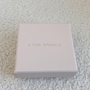 Puede incluir: Una peque&ntilde;a caja cuadrada de color rosa p&aacute;lido con el texto "S FOR SPARKLE" en una fuente delicada de color dorado. La caja est&aacute; sobre una superficie texturizada de color blanquecino.