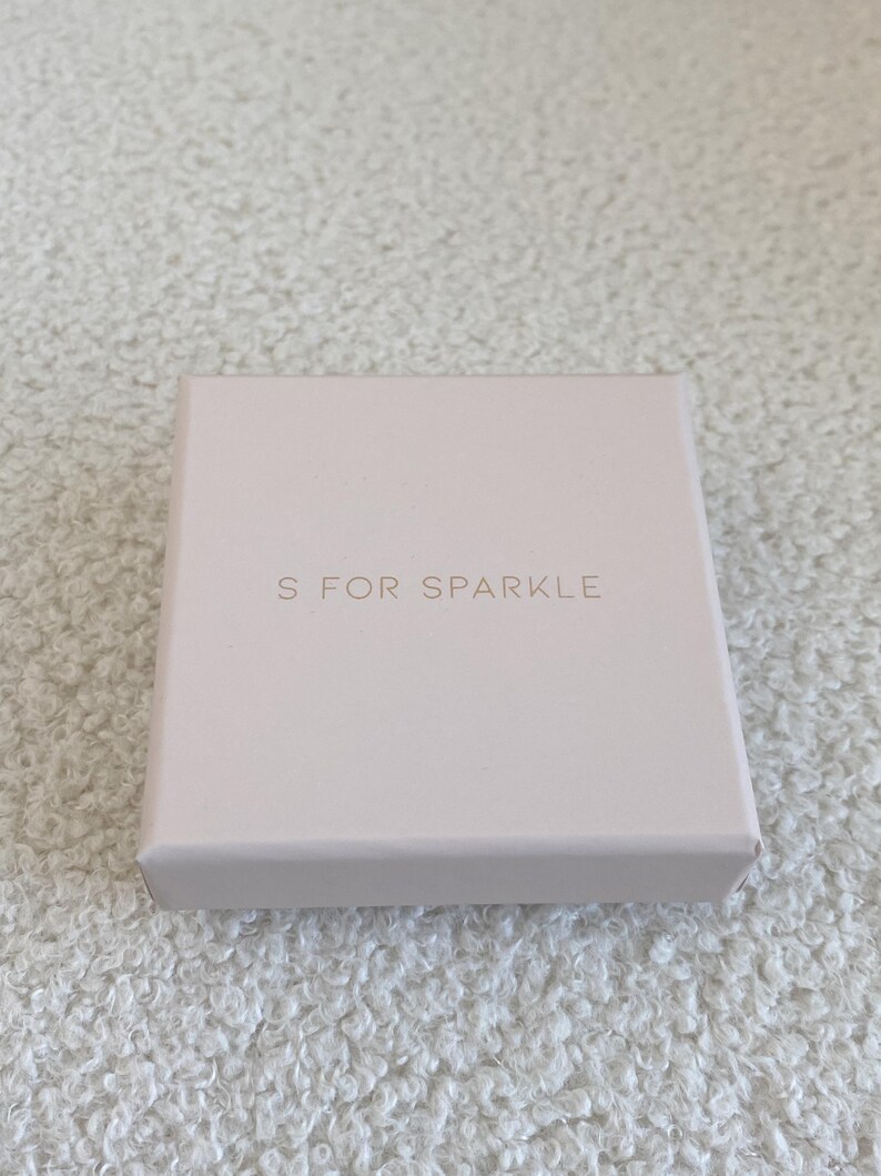 Puede incluir: Una peque&ntilde;a caja cuadrada de color rosa p&aacute;lido con el texto "S FOR SPARKLE" en una fuente delicada de color dorado. La caja est&aacute; sobre una superficie texturizada de color blanquecino.