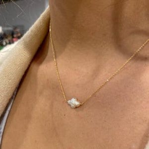 Puede incluir: Un delicado collar de oro con un peque&ntilde;o colgante de perla blanca de forma irregular. El collar se muestra sobre un fondo de tono de piel, destacando el contraste entre la cadena de oro y la perla.