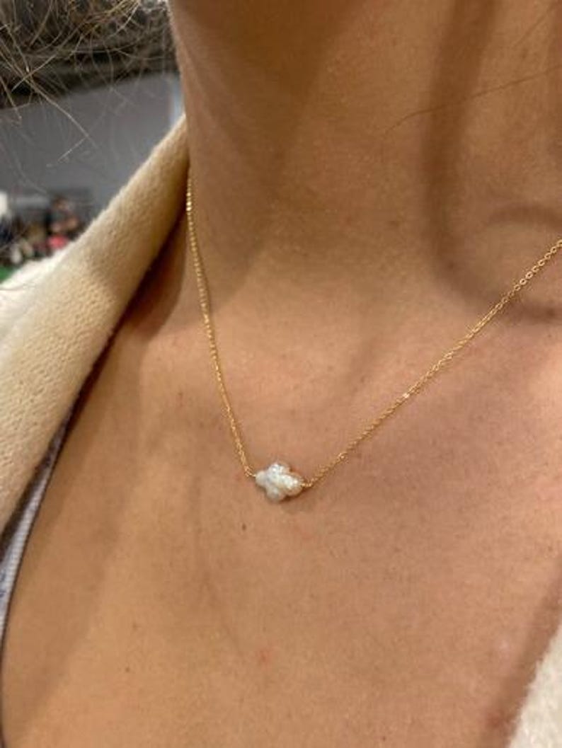Puede incluir: Un delicado collar de oro con un peque&ntilde;o colgante de perla blanca de forma irregular. El collar se muestra sobre un fondo de tono de piel, destacando el contraste entre la cadena de oro y la perla.