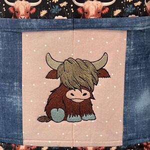May include: A fabric item featuring a Highland cow design. The central panel has an embroidered cartoon cow on a pink background with white polka dots. The sides are denim blue, and the top and bottom have a black floral pattern with cow faces.