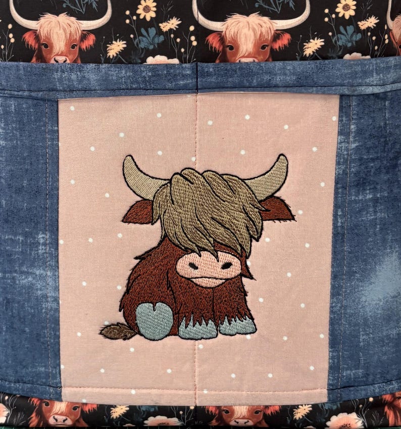 May include: A fabric item featuring a Highland cow design. The central panel has an embroidered cartoon cow on a pink background with white polka dots. The sides are denim blue, and the top and bottom have a black floral pattern with cow faces.