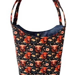May include: A black tote bag with a repeating pattern of Highland cows and flowers in shades of orange, yellow, and white. The bag has a denim blue interior and matching handles.
