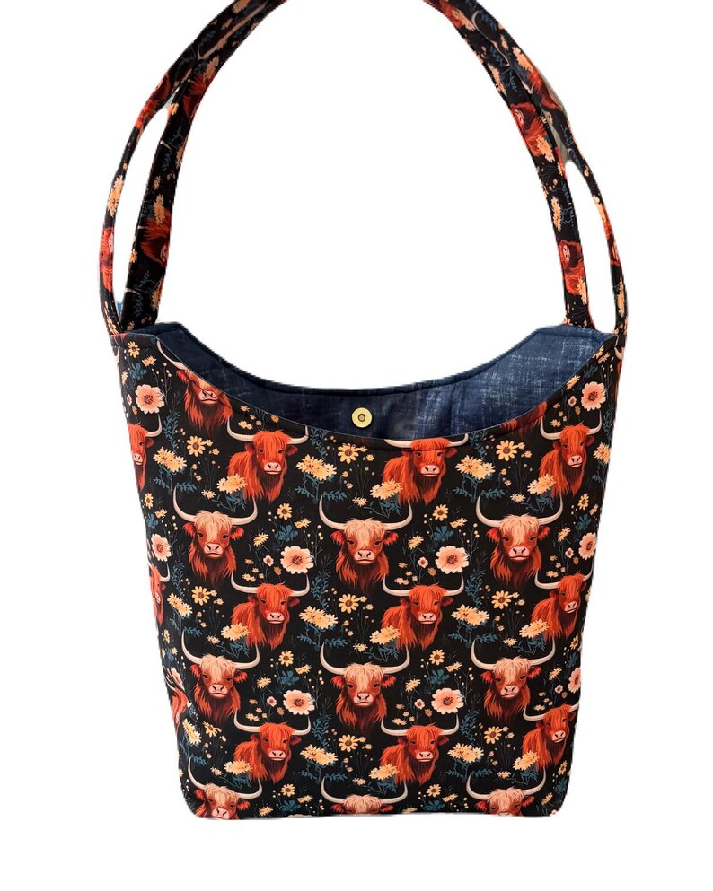 May include: A black tote bag with a repeating pattern of Highland cows and flowers in shades of orange, yellow, and white. The bag has a denim blue interior and matching handles.