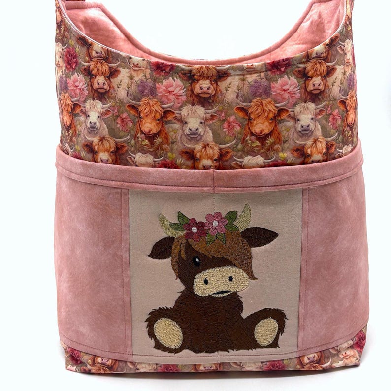 May include: A pink fabric storage basket with a floral cow print and a stitched cow applique. The top and bottom edges feature a cow and floral pattern. The front of the basket has a light tan panel with a brown cow applique wearing a floral crown.