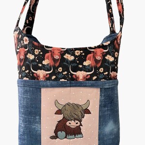 May include: A shoulder bag with a Highland cow theme. The top section features a black background with red-haired Highland cows and white flowers. The bottom is denim blue with a pink square embroidered with a cartoon Highland cow.