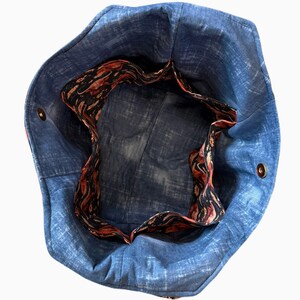 May include: A blue denim tote bag with a patterned interior. The bag has a circular shape with a contrasting interior lining featuring a swirling pattern in shades of red, black, and gold. The bag has two metal snap closures.