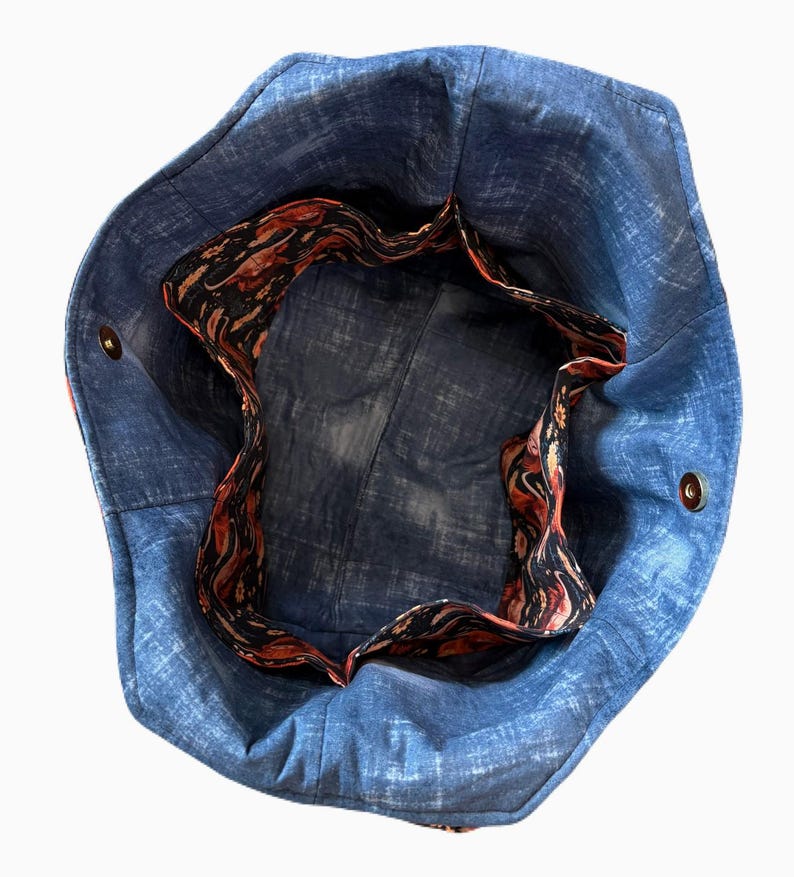 May include: A blue denim tote bag with a patterned interior. The bag has a circular shape with a contrasting interior lining featuring a swirling pattern in shades of red, black, and gold. The bag has two metal snap closures.
